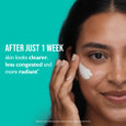 Person applying white exfoliating cream to cheek against turquoise background.