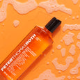 Orange gel cleanser bottle with black cap surrounded by foam and bubbles on orange background.
