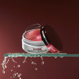 White jar of pink hydra-gel eye patches with black lid on glass rod against burgundy background with water droplets.