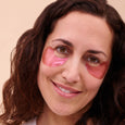 Woman wearing pink hydrogel eye patches under both eyes against a beige background.