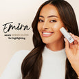White stick highlighter held next to woman's cheekbone, showing subtle luminous finish on skin.