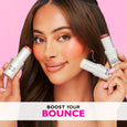 Woman holding two white cylindrical makeup sticks with coral-toned shimmer on cheeks against pink background.